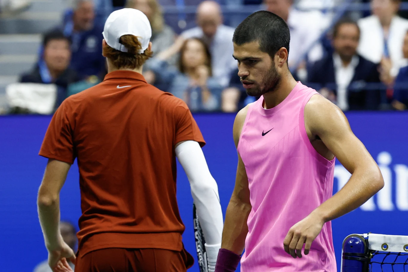 Dwóch tenisistów na korcie podczas turnieju, jeden ubrany w różową koszulkę bez rękawów, drugi w czerwonym stroju i białej czapce, wokół nich widownia uważnie obserwująca przebieg meczu.