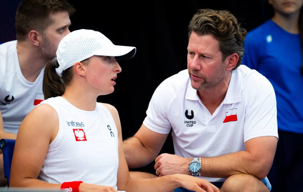 Iga Świątek i Wim Fissette cieszyli się w niedzielę z tego samego powodu - awansu reprezentacji w BJK Cup Rozmowa dwóch osób w sportowych strojach z polskimi emblematami, siedzących wspólnie podczas wydarzenia sportowego. Mężczyzna delikatnie trzyma kobietę za ramię i patrzy na nią, wykazując zainteresowanie oraz wsparcie. W tle trzecia osoba oraz elementy...