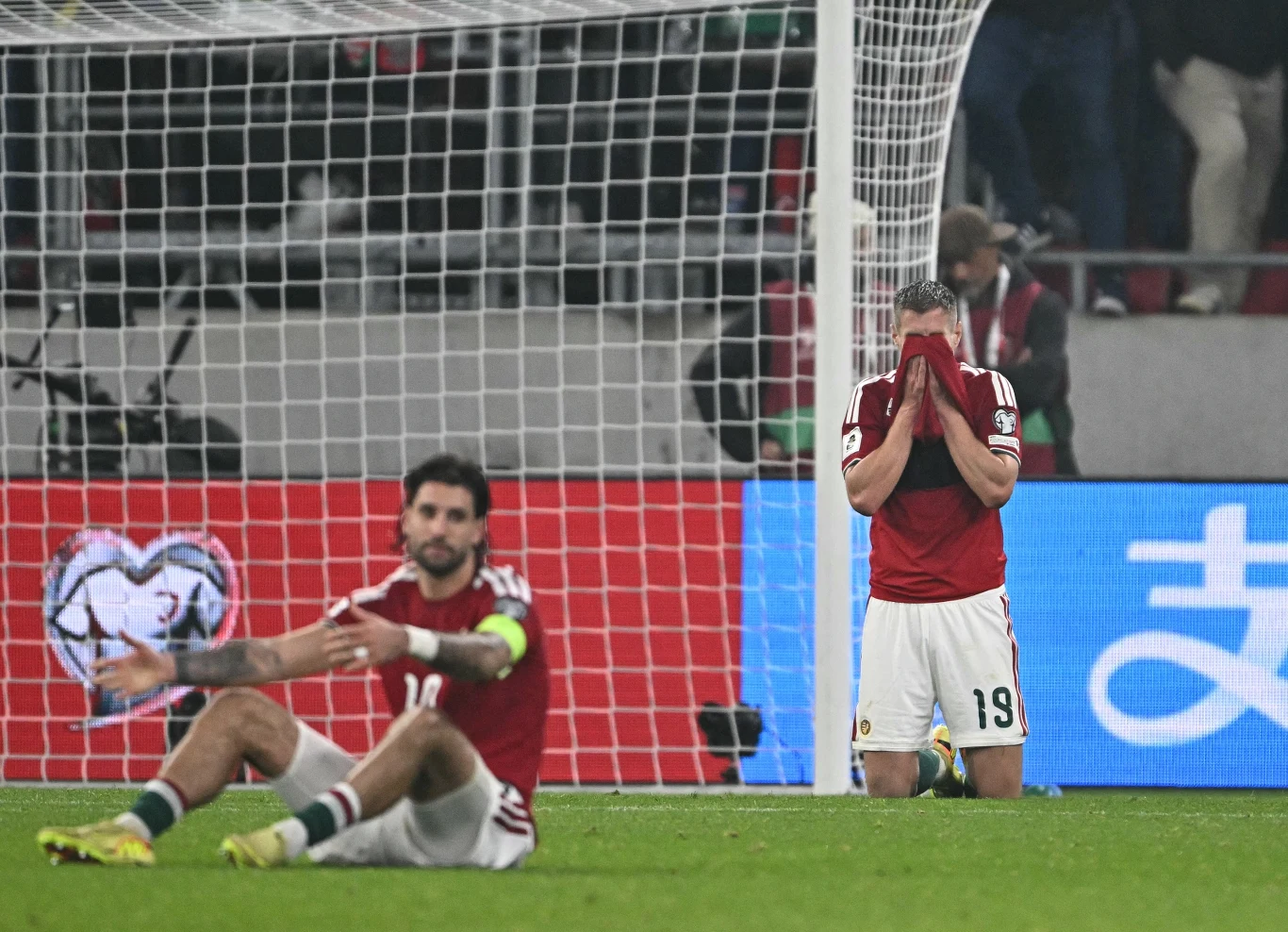 Mundial znowu przesunął się Węgrom na horyzont - dokładnie na 2030 rok... Dwóch piłkarzy w czerwonych koszulkach wyrażających rozczarowanie na tle bramki, jeden z nich siedzi na murawie ze zrezygnowaną miną, drugi stoi z twarzą ukrytą w dłoniach.