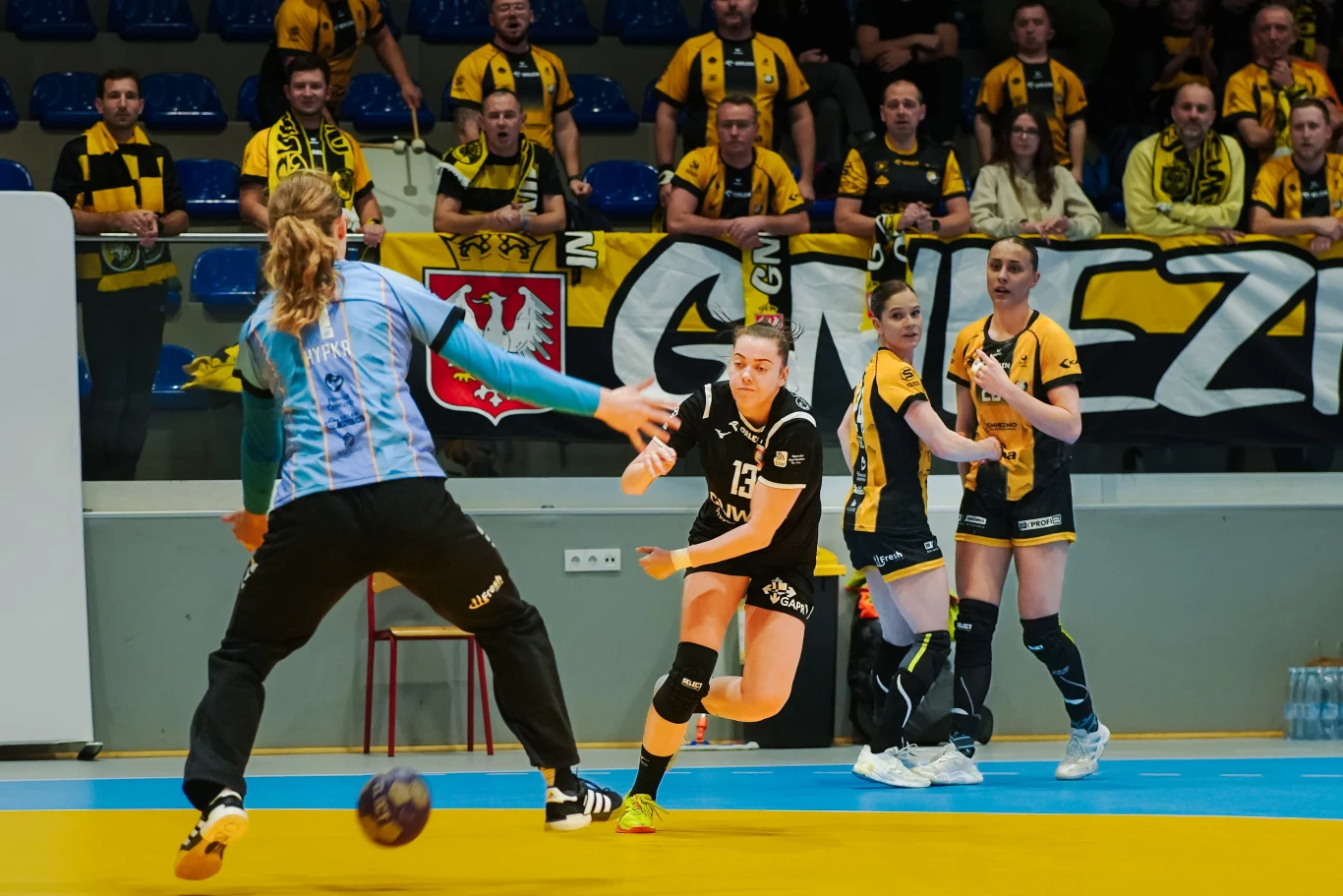 Zawodniczka drużyny piłki ręcznej w czarno-żółtym stroju atakuje bramkę, bramkarka w niebieskim stroju szykuje się do obrony. W tle kibice drużyny w żółto-czarnych barwach oraz wywieszony transparent z napisem Gniezno. Po prawej stronie widoczne trzy z...