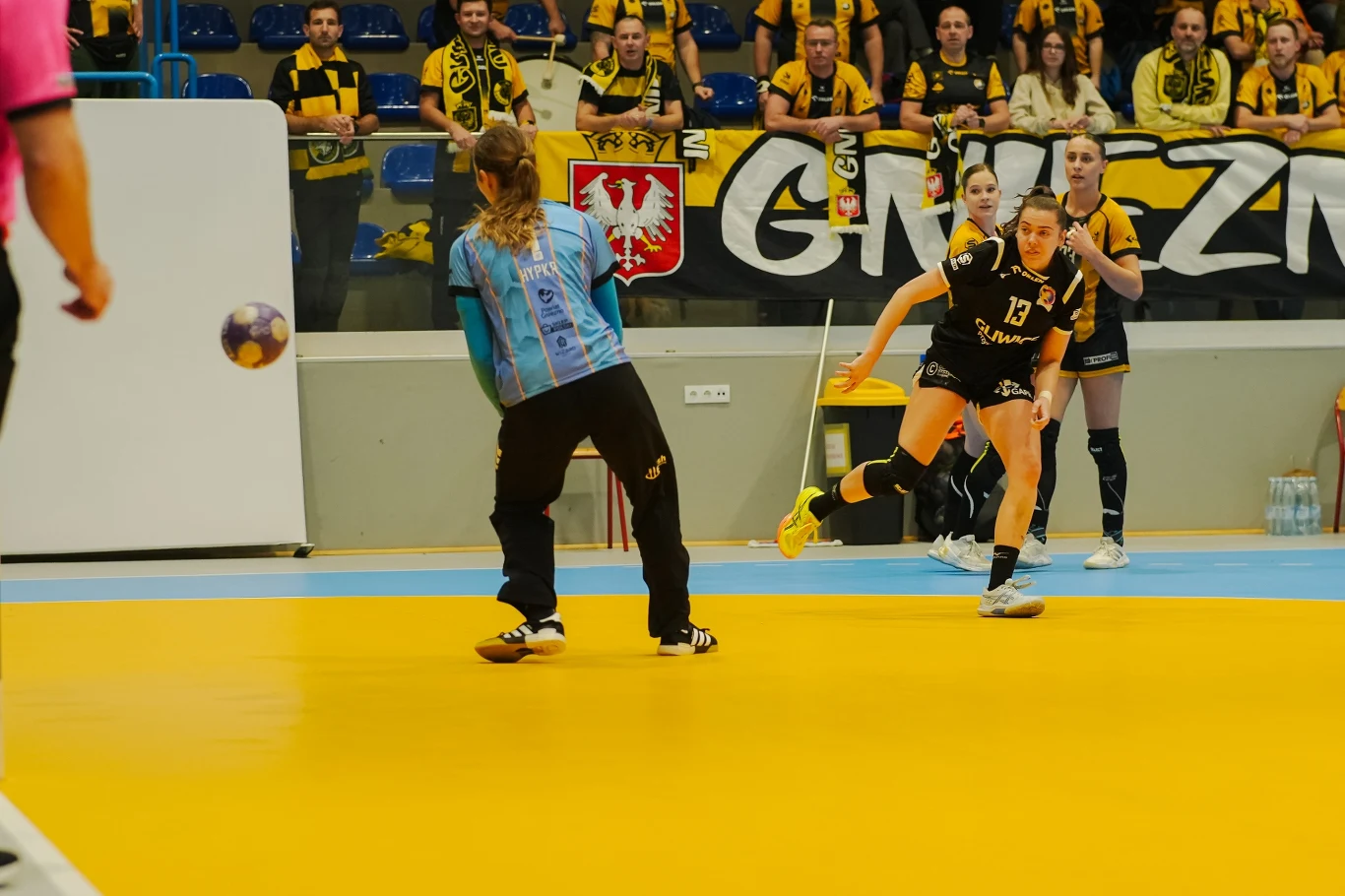 Zawodniczka drużyny w czarnych strojach wykonuje rzut podczas meczu piłki ręcznej, próbując pokonać bramkarkę drużyny przeciwnej ubranej w niebieską koszulkę. Na trybunach kibice dopingują swoją drużynę, trzymając żółto-czarne szaliki i transparenty.