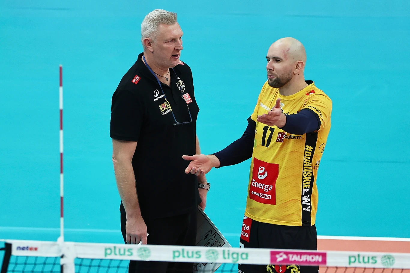 Trener Energa Trefla Gdańsk Mariusz Sordyl i przyjmujący Piotr Orczyk Siatkarz w żółto-czarnej koszulce rozmawia z trenerem na boisku do siatkówki, obaj stoją po jednej stronie siatki, otoczeni sportowym wyposażeniem hali.