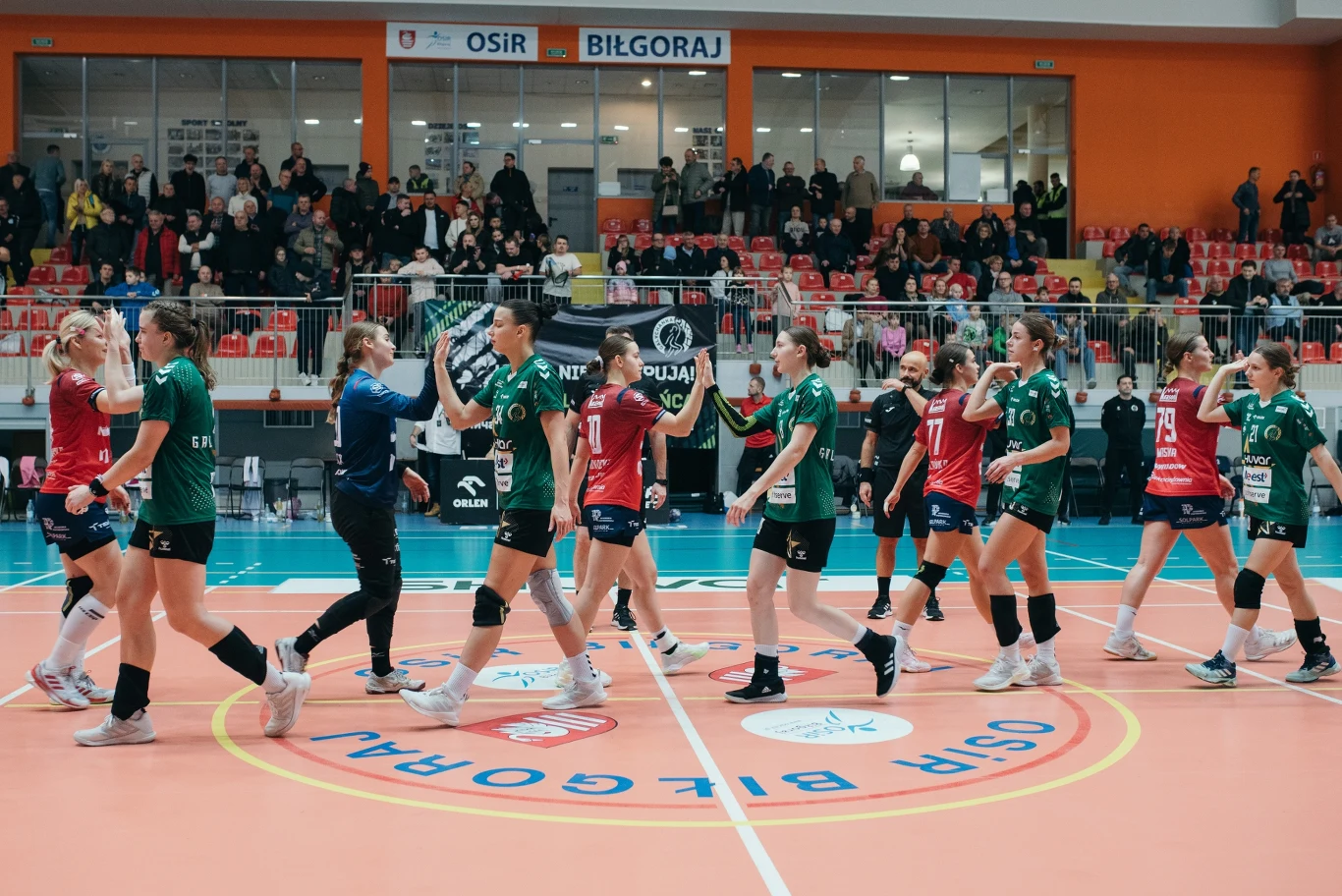 Dwie drużyny kobiecego zespołu piłki siatkowej witają się na środku hali sportowej, zawodniczki ubrane w zielone i czerwone stroje, wokół pełne trybuny z kibicami oraz widoczne bandy reklamowe.