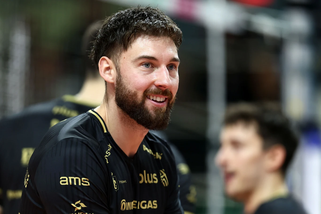Sportowiec z brodą i krótkimi włosami ubrany w czarny koszulkę z logo sponsora i nazwą drużyny siatkarskiej, uśmiechnięty, w tle inny zawodnik lekko rozmyty.