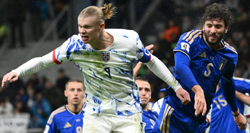 Erling Haaland (na pierwszym planie) to już dziś mocny kandydat na króla strzelców mundialu Dwóch piłkarzy rywalizuje o piłkę podczas meczu, jeden w białej koszulce z niebieskimi wzorami i opaską kapitana, drugi w niebieskim stroju sportowym, wokół nich widoczny stadion i kibice.