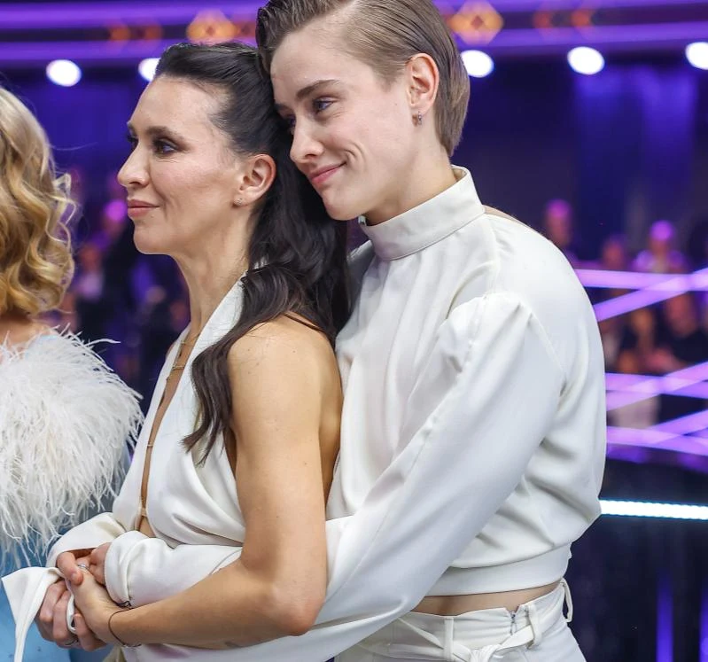 Katarzyna Zillmann i Janja Lesar Dwie kobiety ubrane w eleganckie, jasne stroje stoją blisko siebie, jedna obejmuje drugą od tyłu z uśmiechem na twarzy, w tle rozmyte światła i sylwetki innych osób.