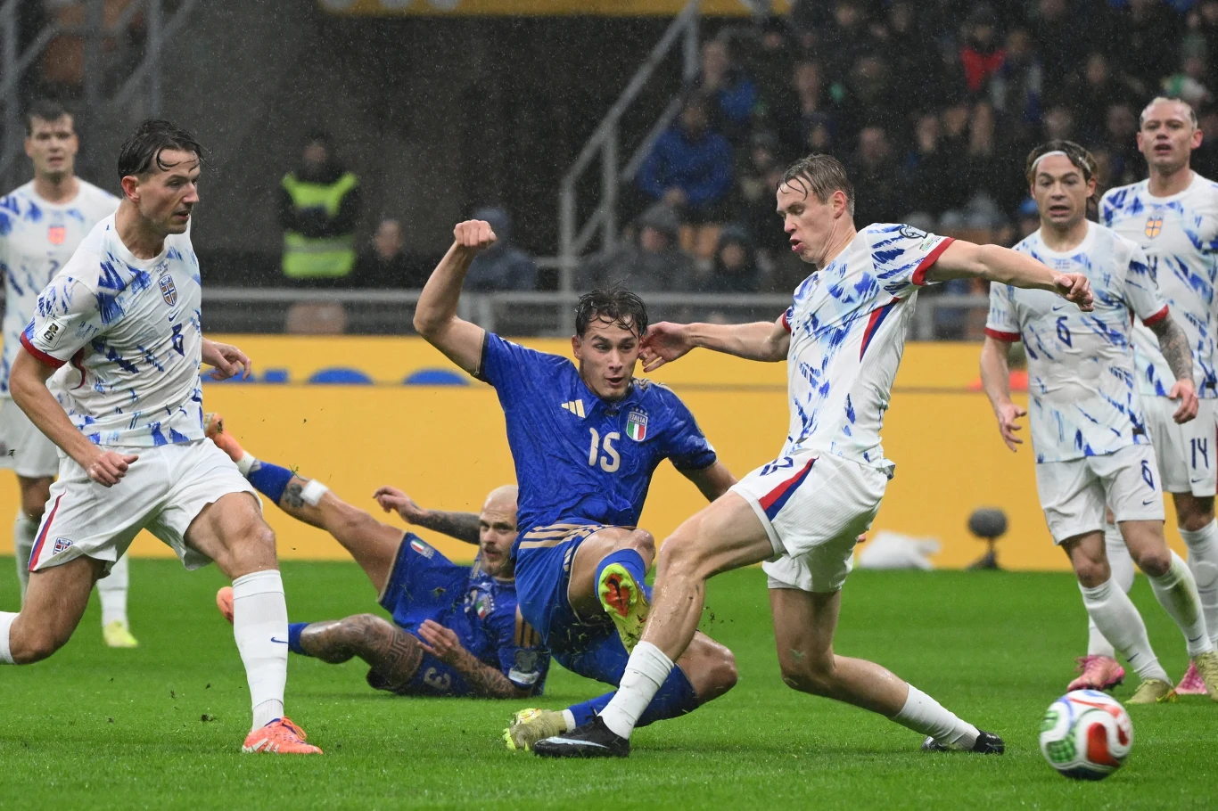 Dynamiczna walka piłkarzy podczas meczu, zawodnik w niebieskim stroju próbuje przejąć piłkę od rywali w białych strojach z niebiesko-czerwonymi akcentami, w tle widoczna murawa i kibice.