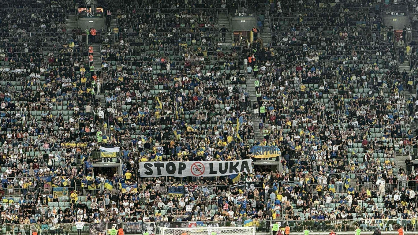 Tłum kibiców na trybunach stadionu trzymający transparent z napisem STOP LUKA, otoczony różnorodnymi flagami i szalikami klubowymi.