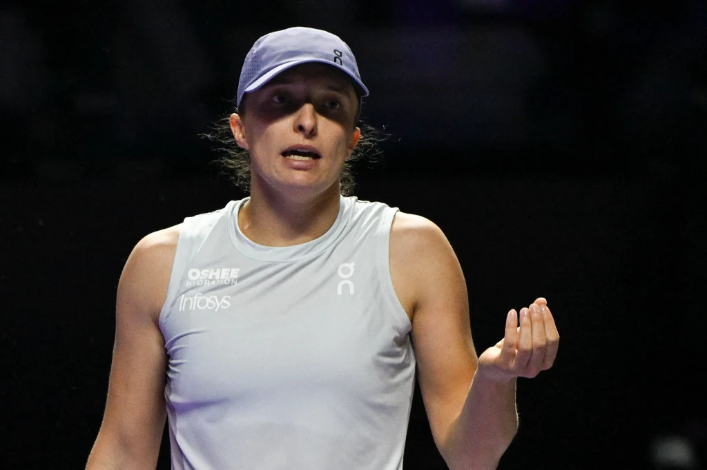 Iga Świątek Kobieta w sportowym stroju i czapce z daszkiem podczas meczu tenisa, w dynamicznej pozie, gestykuluje ręką, na tle ciemnego rozmytego otoczenia. Na jej koszulce widoczne są logotypy sponsorów.