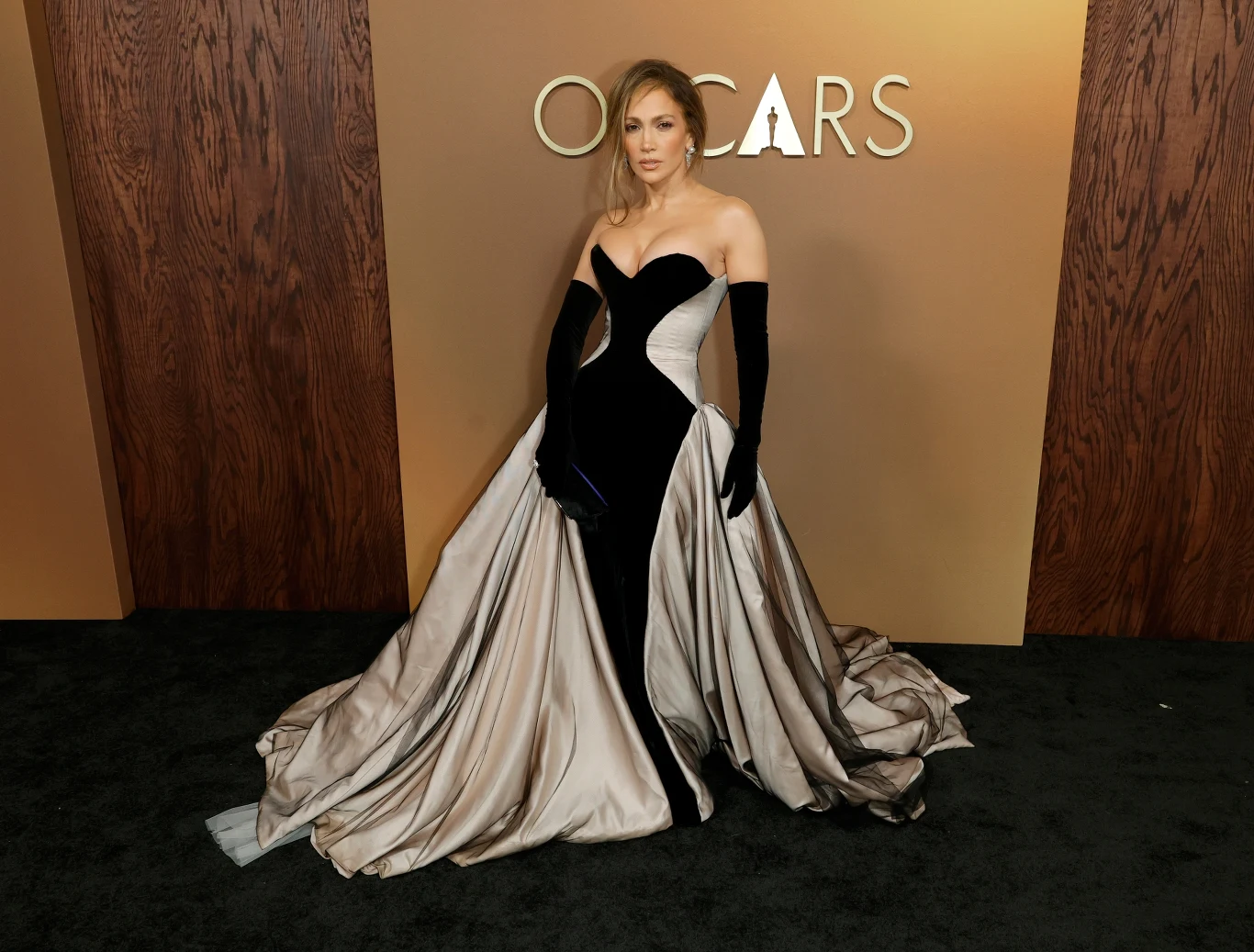 Kobieta w efektownej, eleganckiej sukni z czarno-białym gradientem, stojąca na tle ściany z napisem OSCARS. Stylizacja uzupełniona długimi czarnymi rękawiczkami i wyrazistą fryzurą.