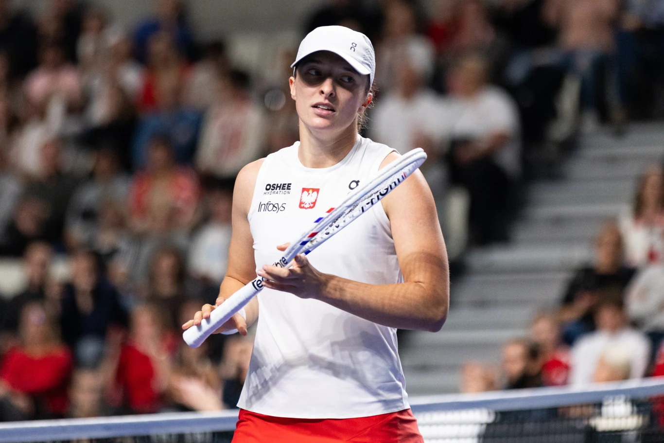 Tenisistka w białej koszulce oraz czapce trzymająca rakietę tenisową, stojąca na korcie podczas meczu, w tle rozmazana publiczność na trybunach.