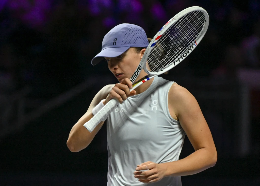 Tenisistka w sportowym stroju i czapce, trzymająca rakietę tenisową oraz delikatnie dotykająca ją twarzą, ujęcie z kortu podczas meczu przy mocnym świetle punktowym.