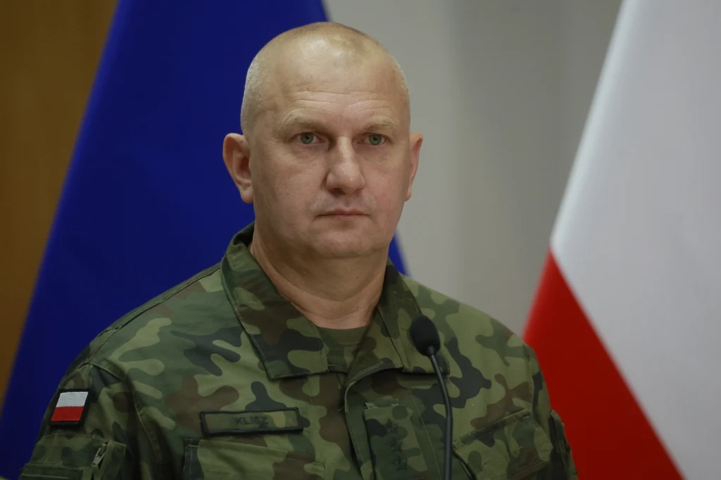 Gen. Maciej Klisz Mężczyzna w mundurze wojskowym stoi przed mikrofonem, w tle widoczne flagi Polski oraz Unii Europejskiej.