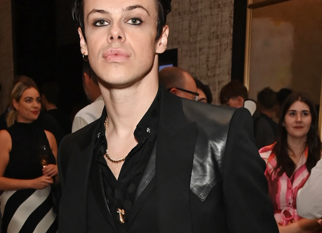 Yungblud odwołał resztę swoich występów ze względu na zdrowie Mężczyzna w czarnym garniturze i makijażu z mocno podkreślonymi oczami stoi w eleganckim wnętrzu, w tle widoczne są inne osoby ubrane w wieczorowe stroje.