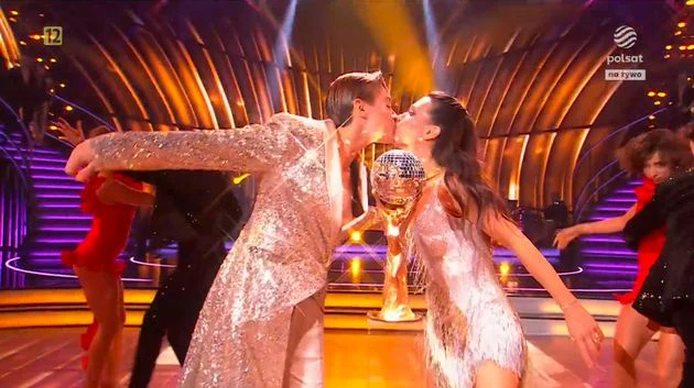 Dwie tancerki ubrane w połyskujące kostiumy całują się na środku sceny z podniesionym pucharem w kształcie kuli lustrzanej, w tle pozostałe osoby i scenografia programu rozrywkowego telewizyjnego.