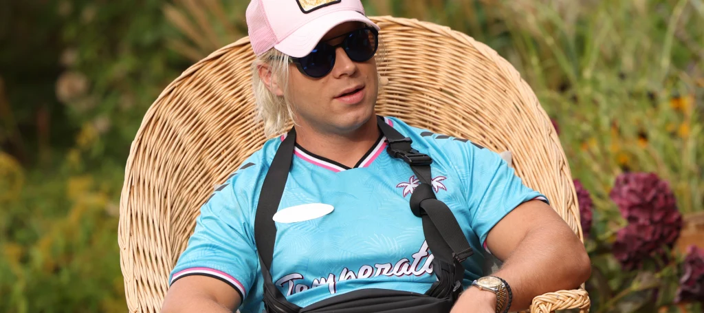 Na posesję Skolima wtargnęło trzech mężczyzn Mężczyzna w jasnoniebieskiej koszulce sportowej, różowej czapce z daszkiem i okularach przeciwsłonecznych siedzi w wiklinowym fotelu na tle roślin ogrodowych. Ma założony temblak na prawą rękę.