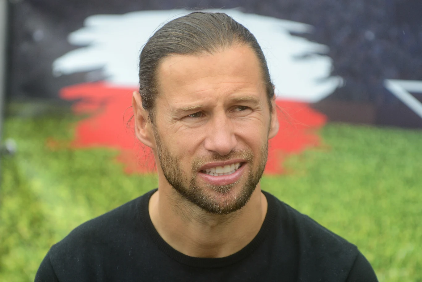 Grzegorz Krychowiak Mężczyzna z długimi włosami spiętymi do tyłu oraz krótką brodą, uśmiechający się lekko, w tle rozmyte barwy przypominające polską flagę i zielone tło.