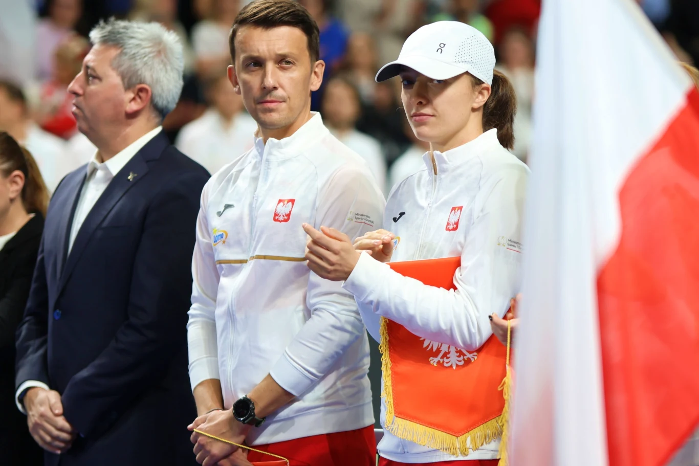 Dwie osoby w sportowych ubraniach z godłem Polski stoją obok siebie na tle flagi Polski, obok nich znajduje się mężczyzna w garniturze, w tle widoczne są rozmazane sylwetki innych uczestników wydarzenia.