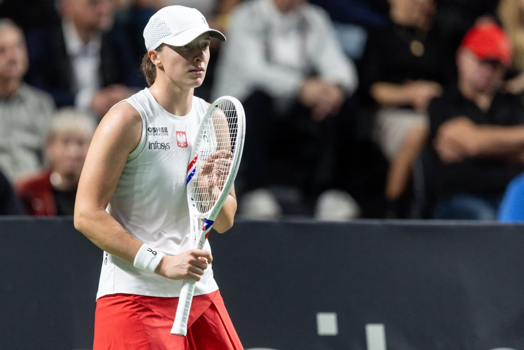 Tenisistka w białej czapce i stroju sportowym trzyma rakietę tenisową na tle trybun pełnych widzów.