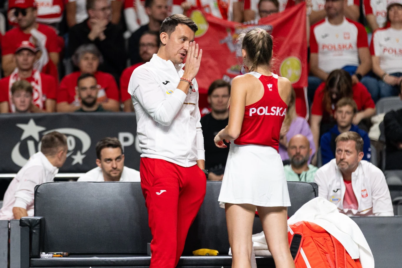 Trener w białej bluzie i czerwonych spodniach rozmawia z zawodniczką reprezentacji Polski w czerwono-białym stroju tenisowym, w tle widoczni kibice w biało-czerwonych barwach oraz polskie flagi.
