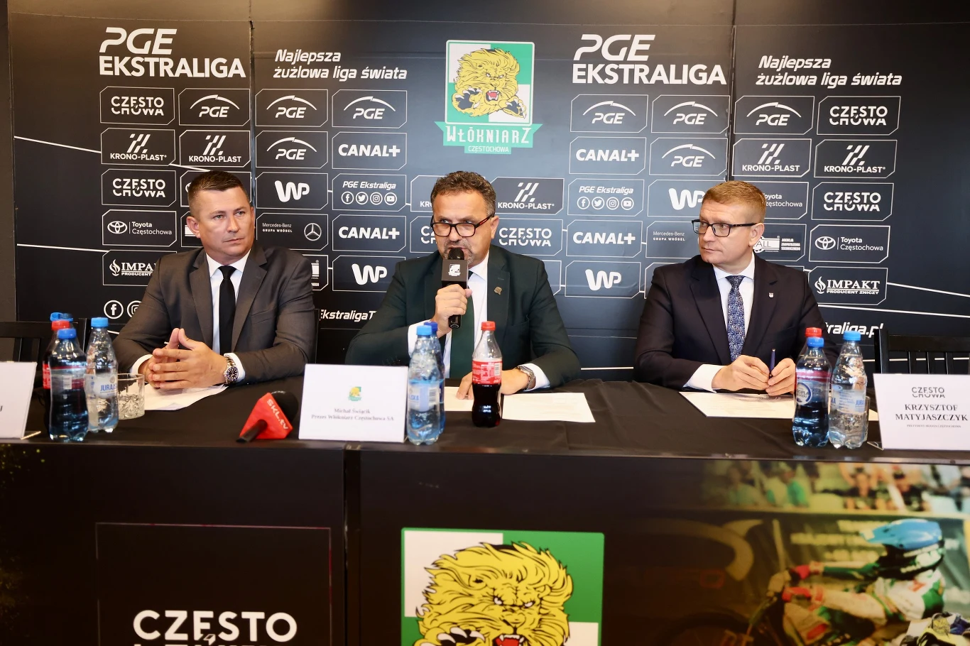 Trzech mężczyzn w garniturach siedzi za stołem na tle tablicy z logotypami sponsorów oraz klubów żużlowych. Środkowy mężczyzna mówi do mikrofonu, przed nimi stoją butelki wody, szklanki i karteczki z nazwiskami. Po lewej stronie widoczny jest herb klub...