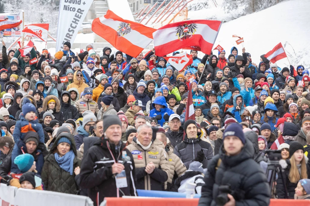 Kibice na torze Igls koło Innsbrucka Tłum kibiców ubranych w zimowe ubrania na tle zaśnieżonego otoczenia, wiele austriackich flag uniesionych w powietrzu, różnorodność wiekowa widzów, część z nich trzyma aparaty fotograficzne i kamery, atmosfera radosna i pełna entuzjazmu.