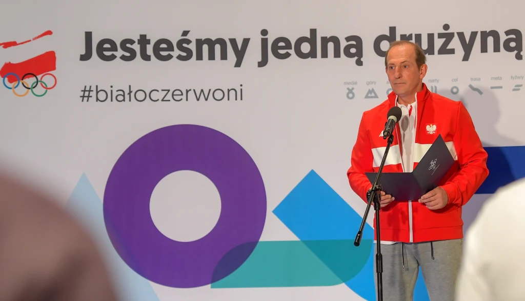 Marek Skowroński Mężczyzna w biało-czerwonym dresie z emblematem Polski przemawia przy mikrofonie, trzymając dokument, na tle ścianki z napisem Jesteśmy jedną drużyną i olimpijskimi symbolami.