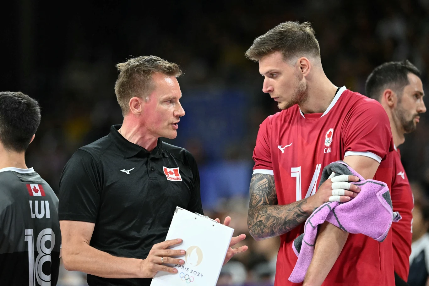 Arthur Szwarc w reprezentacji Kanady Trener rozmawia z zawodnikiem ubranym w czerwony strój, trzymającym ręcznik na ramieniu. W tle widać innych członków drużyny oraz halę sportową podczas meczu siatkówki.