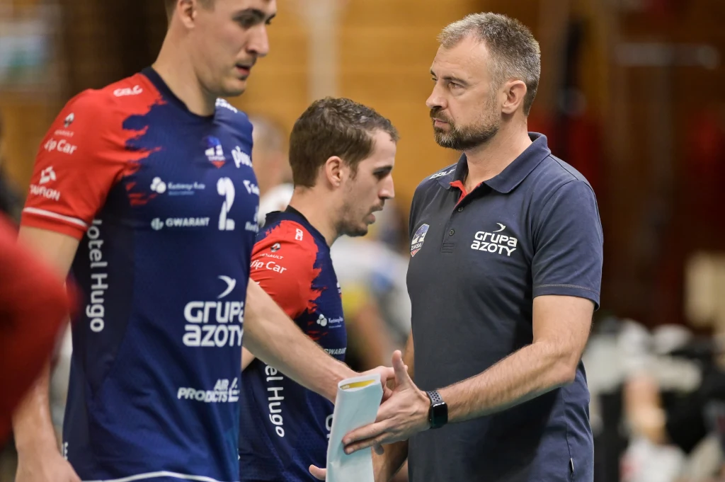 Nikola Grbić jako szkoleniowiec ZAKSY Kędzierzyn-Koźle Trener drużyny siatkarskiej w granatowej koszulce z logo Grupa Azoty rozmawia z zawodnikami podczas przerwy w meczu, w tle widoczni sportowcy w strojach sportowych.