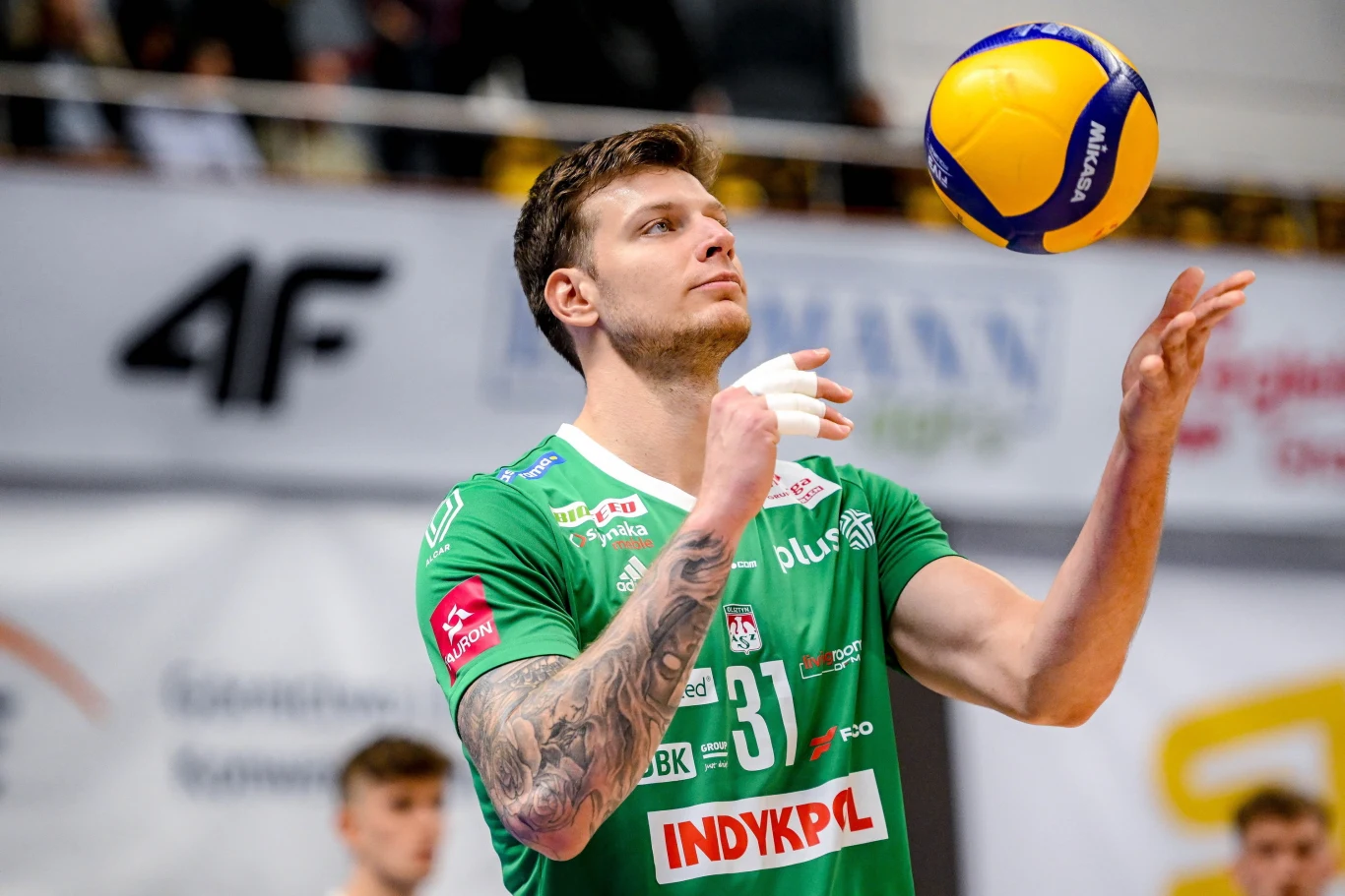 Athur Szwarc w barwach Indykpol AZS-u Olsztyn Siatkarz w zielonym stroju drużynowym z numerem 31 skupia się na piłce do siatkówki, którą zamierza podać. W tle widoczni są kibice oraz elementy hali sportowej.