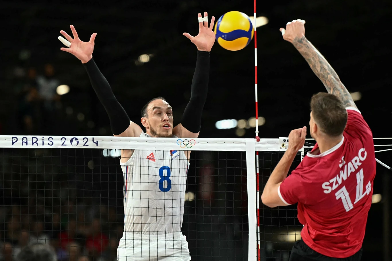 Arthur Szwarc w reprezentacji Kanady Dwóch siatkarzy podczas dynamicznej akcji na boisku. Zawodnik w białym stroju próbuje zablokować piłkę nad siatką, podczas gdy zawodnik w czerwonej koszulce atakuje. Nad siatką widoczny napis Paris 2024 oraz olimpijskie kółka.