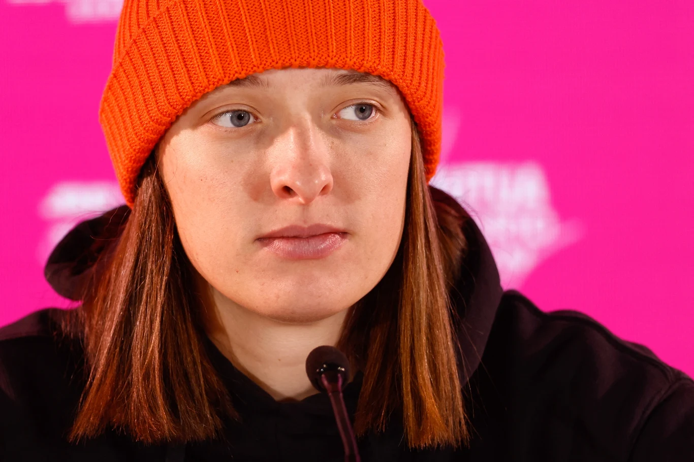 Iga Świątek Osoba w pomarańczowej czapce i czarnej bluzie siedzi przed mikrofonem na tle różowego ekranu.
