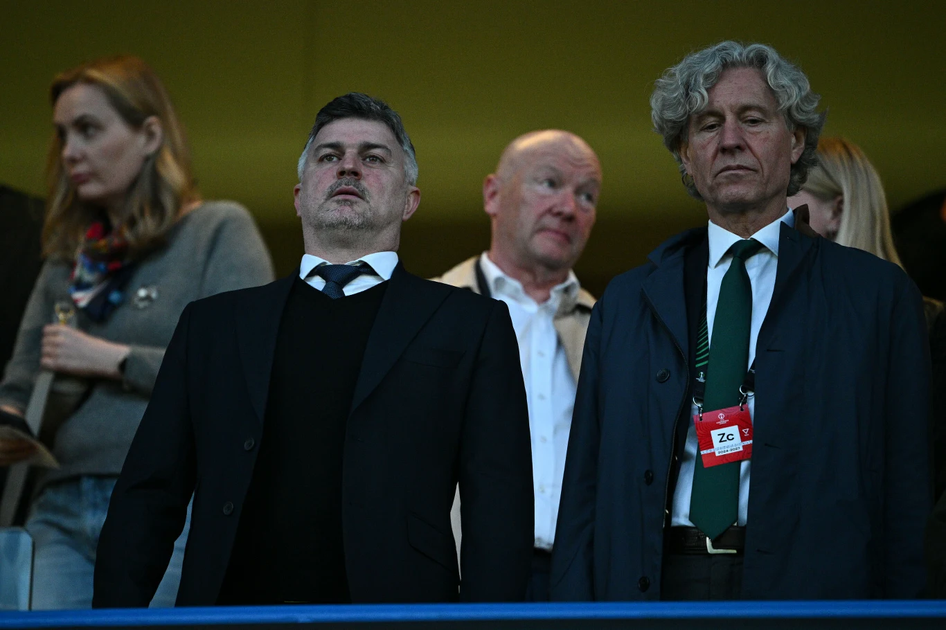 Michał Żewłakow i Dariusz Mioduski Grupa czterech osób na trybunach, dwaj mężczyźni na pierwszym planie ubrani elegancko, jeden z nich z identyfikatorem na szyi, w tle dwie osoby obserwujące wydarzenie.