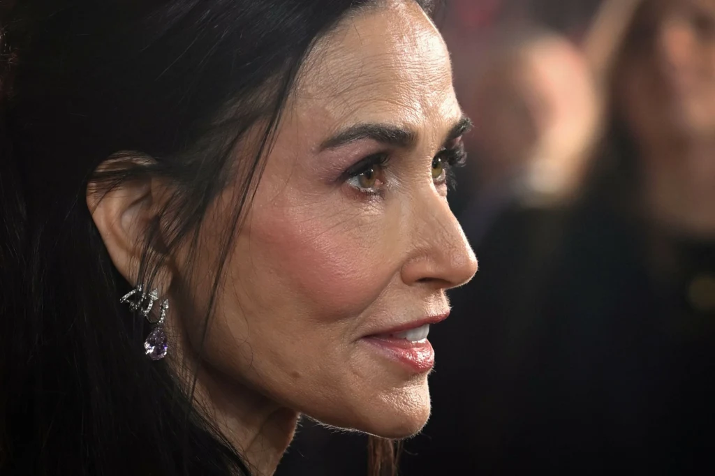 Zbliżenie kobiecej twarzy z długimi, ciemnymi włosami, eleganckimi kolczykami i delikatnym makijażem, widoczna jest spokojna, lekko uśmiechnięta mina oraz światło podkreślające rysy twarzy.