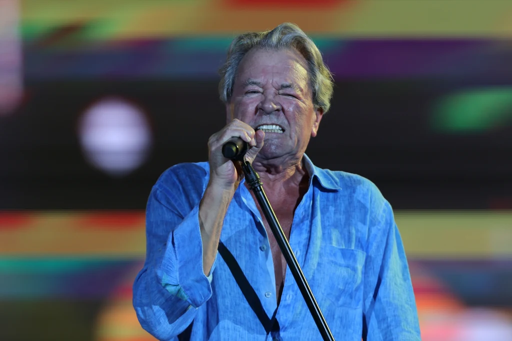 Ian Gillan jest wokalistą Deep Purple Starszy mężczyzna w niebieskiej koszuli śpiewa do mikrofonu na tle kolorowych, rozmytych świateł scenicznych. Jego twarz wyraża silne emocje.