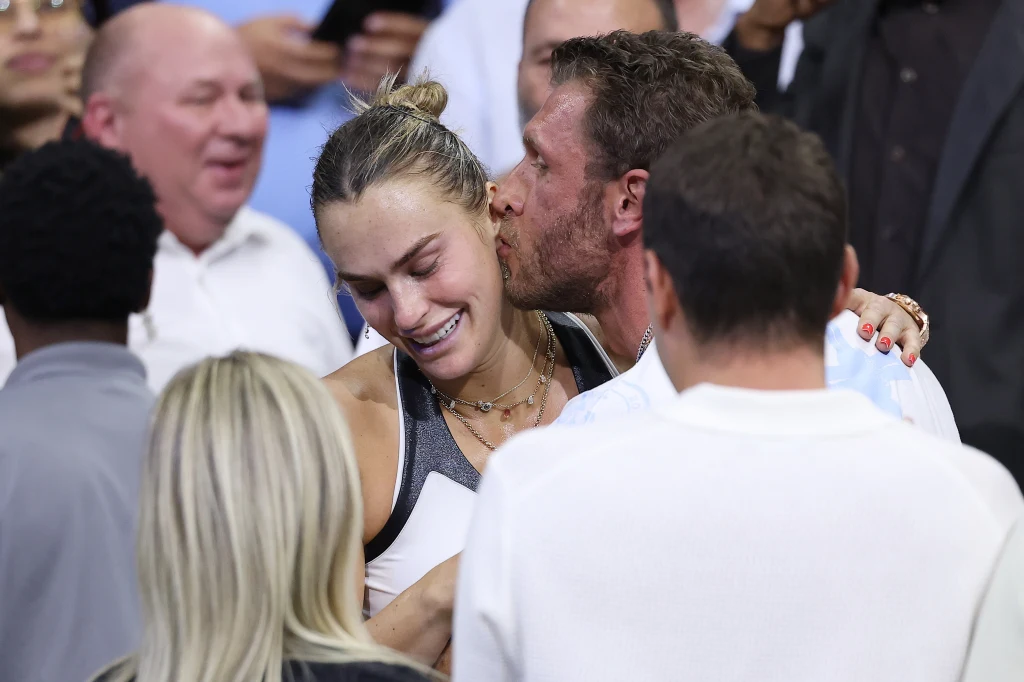 Aryna Sabalenka i Georgios Frangulis Sportowczyni w stroju sportowym uśmiecha się i otrzymuje pocałunek w policzek od mężczyzny w otoczeniu innych osób, atmosfera radości i wsparcia po osiągnięciu sukcesu.
