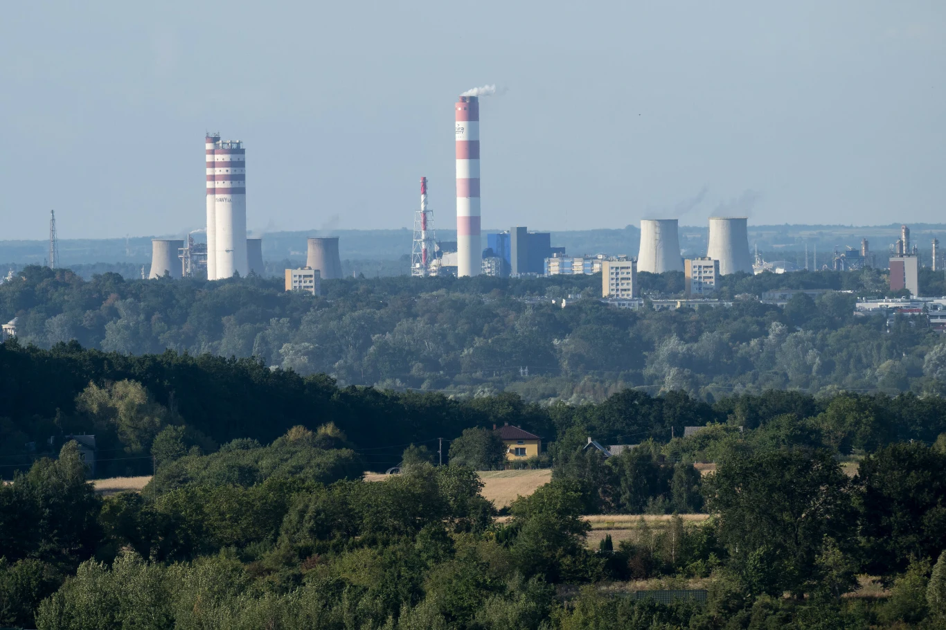 Widok na zakłady azotowe w Puławach Kompleks przemysłowy z wysokimi kominami i chłodniami kominowymi w tle, otoczony przez gęste lasy oraz rozproszoną zabudowę na pierwszym planie, unoszące się dymy wskazują na aktywną działalność zakładu.
