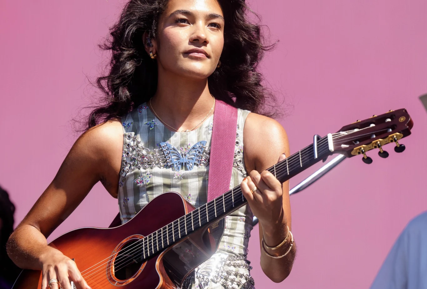 Olivia Dean wystąpi na Orange Warsaw Festivalu! Młoda kobieta z długimi kręconymi włosami grająca na gitarze akustycznej na tle różowej ściany, uśmiechnięta i skupiona, odziana w elegancką, błyszczącą bluzkę bez rękawów.