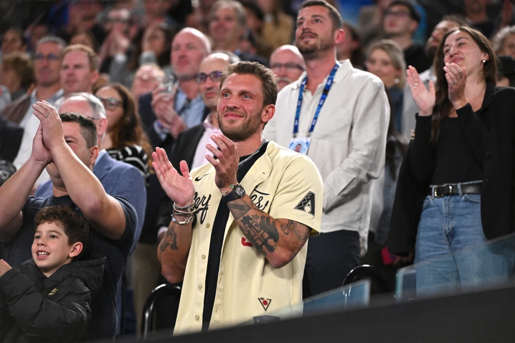 Georgios Frangulis Tłum ludzi na trybunach, większość z nich stoi i klaszcze. Na pierwszym planie mężczyzna w beżowej koszuli, z tatuażami na rękach, uśmiecha się podczas oklasków, obok niego młody chłopiec macha.