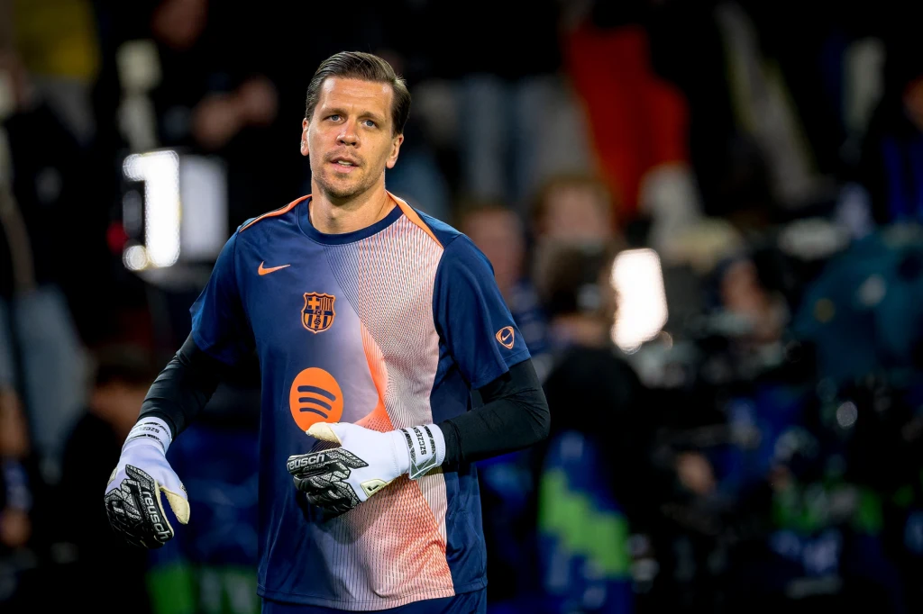 Piłkarz w stroju bramkarza trzyma rękawice na tle boiska. Ubrany w koszulkę z logo klubu piłkarskiego, wokół intensywne oświetlenie i rozmyte sylwetki innych osób.