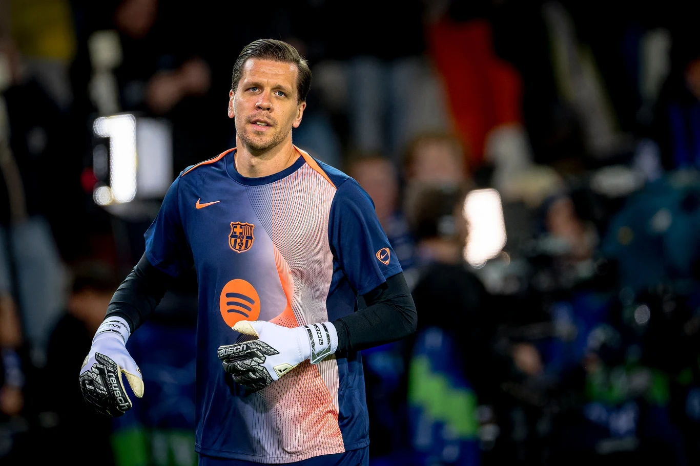 Wojciech Szczęsny Piłkarz w stroju bramkarza trzyma rękawice na tle boiska. Ubrany w koszulkę z logo klubu piłkarskiego, wokół intensywne oświetlenie i rozmyte sylwetki innych osób.