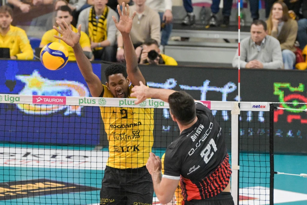 Dwóch siatkarzy w trakcie dynamicznej akcji podczas meczu, zawodnik w żółtym bloku próbuje zatrzymać atak rywala w czarno-czerwonym stroju, widoczne elementy siatki, piłki oraz kibice na trybunach.