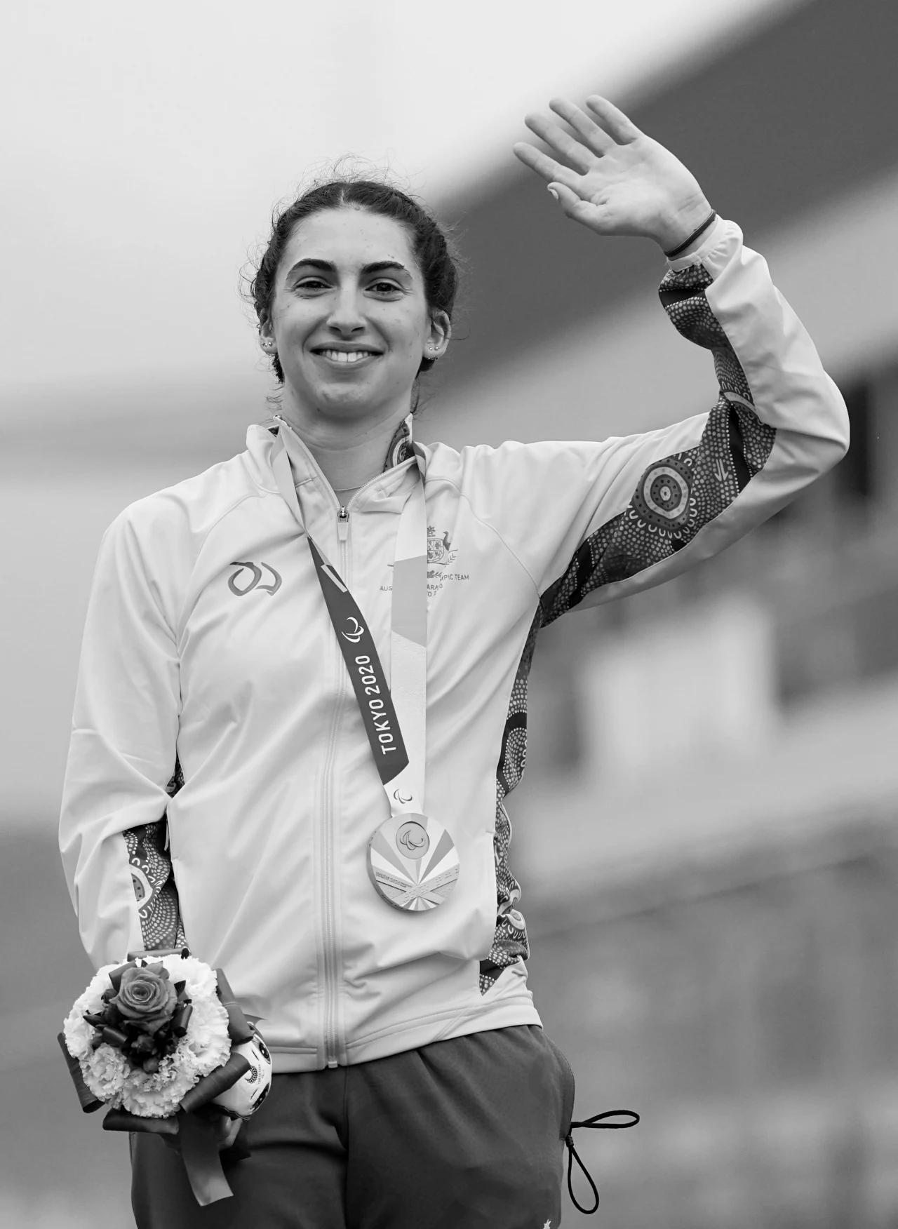 Paige Greco z medalem igrzysk paralimpijskich Uśmiechnięta sportsmenka w dresie z medalem olimpijskim na szyi, trzyma w dłoni bukiet kwiatów i macha ręką w geście pozdrowienia.