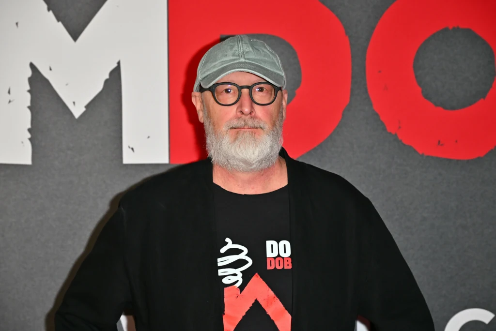 Nowy film Smarzowskiego spotkał się ze skrajnymi emocjami. Wielu zarzuca mu zbyt dużo brutalności Mężczyzna z siwą brodą i w okularach, ubrany w czarną kurtkę oraz czapkę z daszkiem, stoi na tle szarej ściany z dużymi czerwonymi i białymi literami.