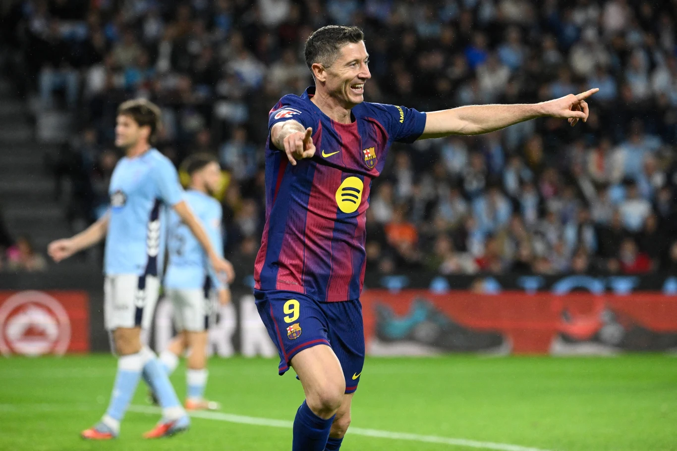 Piłkarz FC Barcelony w stroju meczowym świętuje strzelenie gola na boisku, wskazując rękami w kierunku trybun, w tle zawodnicy drużyny przeciwnej oraz rozmyte postaci na widowni.