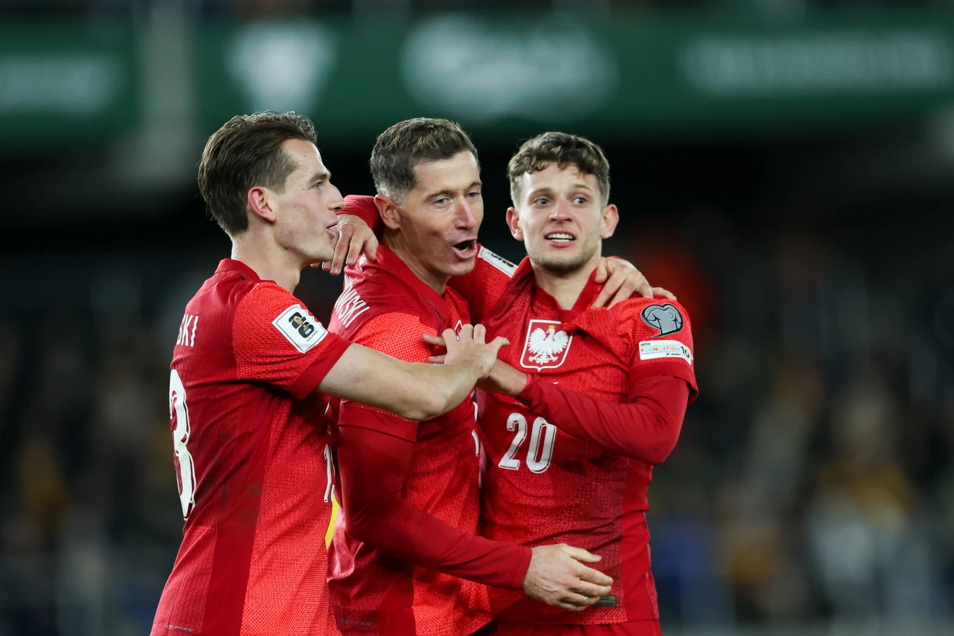 Trzech piłkarzy reprezentacji Polski ubranych w czerwone stroje, obejmujących się wzajemnie na boisku po zdobyciu bramki. W tle widać rozmytą trybunę stadionu.