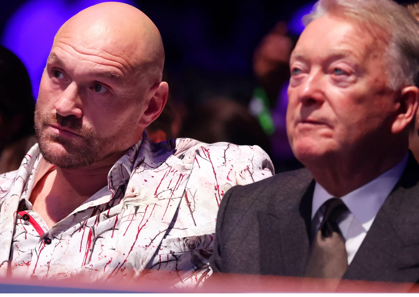 Tyson Fury i Frank Warren, 25.10.2025 r. Dwóch mężczyzn siedzących obok siebie, pierwszy z nich ubrany w jasną koszulę z kolorowym wzorem, drugi w elegancki garnitur z krawatem, obaj patrzą w jednym kierunku z poważnym wyrazem twarzy, tło rozmyte i ciemne, oświetlenie skupia się na postaciach.