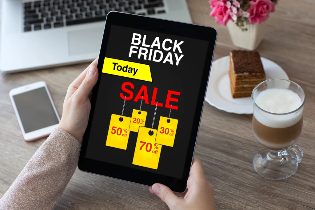 Tablet z widocznym ekranem promującym wyprzedaż Black Friday z dużym napisem SALE, jasno oznaczonymi zniżkami procentowymi oraz tłem z kawą, ciastkiem, laptopem oraz widoczną ręką trzymającą urządzenie.