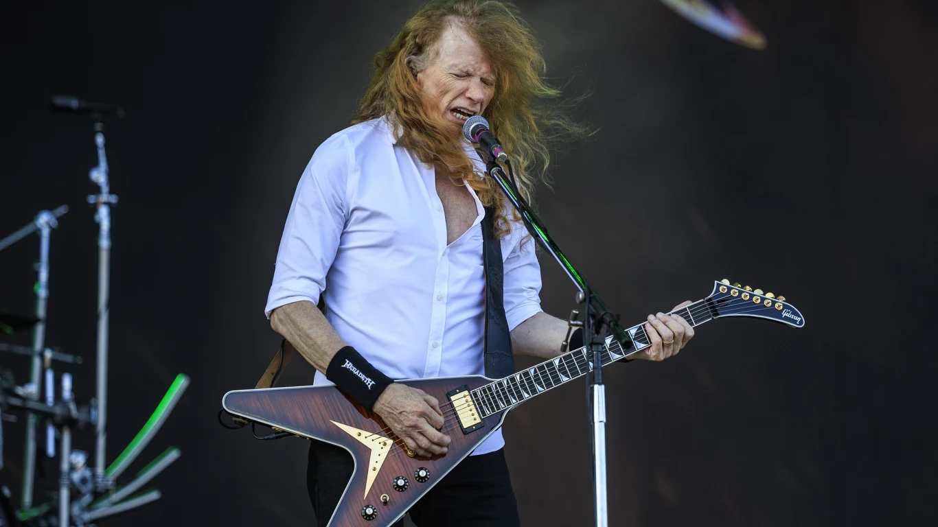 Dave Mustaine (Megadeth) Mężczyzna z długimi rudymi włosami gra na elektrycznej gitarze typu Flying V i śpiewa do mikrofonu na scenie koncertowej, ubrany w białą koszulę z podwiniętymi rękawami, w tle widoczne są elementy perkusji.