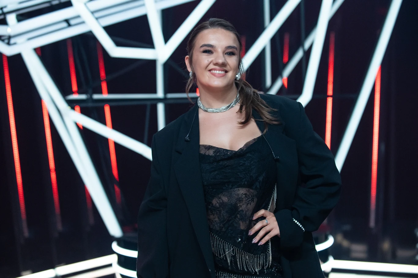 Julia Wasielewska odpadła z "The Voice of Poland" Uśmiechnięta kobieta w eleganckim, czarnym stroju, na tle futurystycznej scenografii z geometrycznymi liniami świetlnymi.